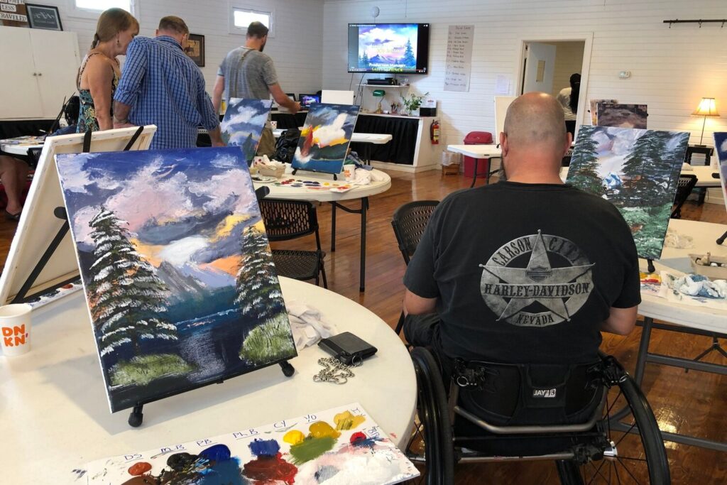 Three male veterans and a female veteran participating in the Healing Art class with a nature painting next to the male veteran in the wheelchair.