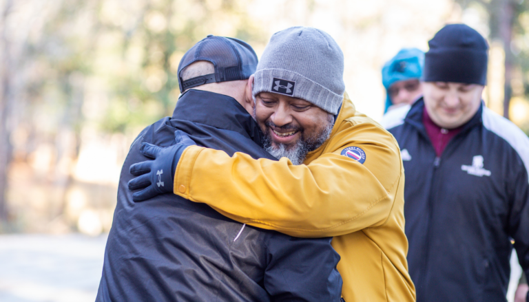 Two men hugging each other. One is wearing a yellow jacket with a grey beanie. The other one is wearing a navy jacket with a hat.