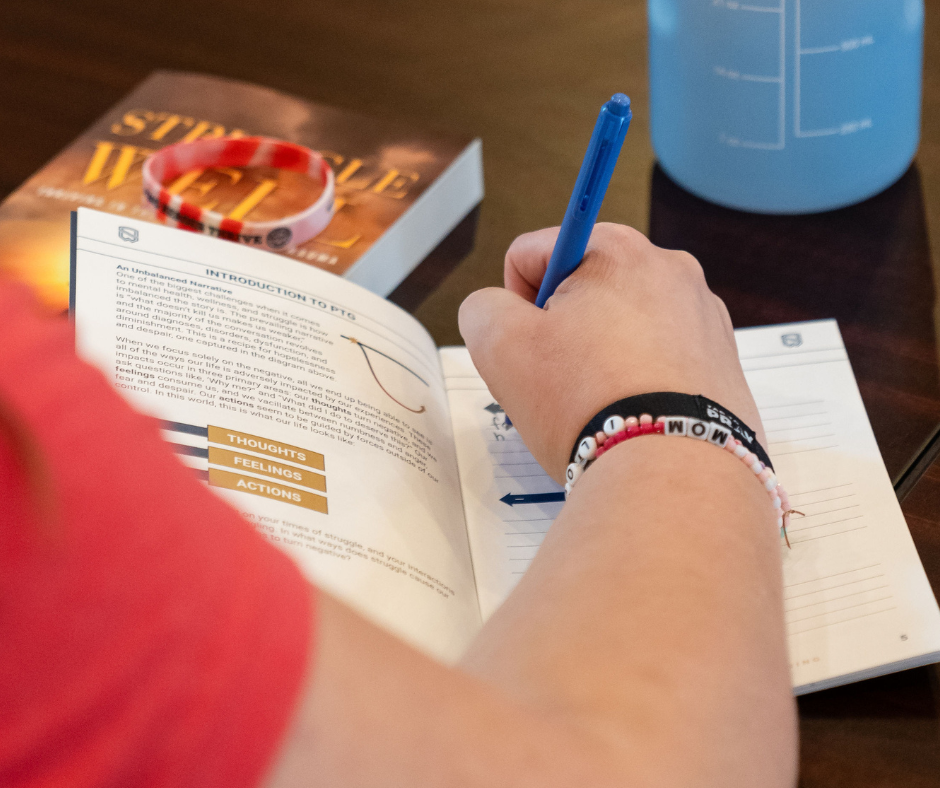 A person writes in a workbook at a table with a book, wristbands, and a water bottle nearby.