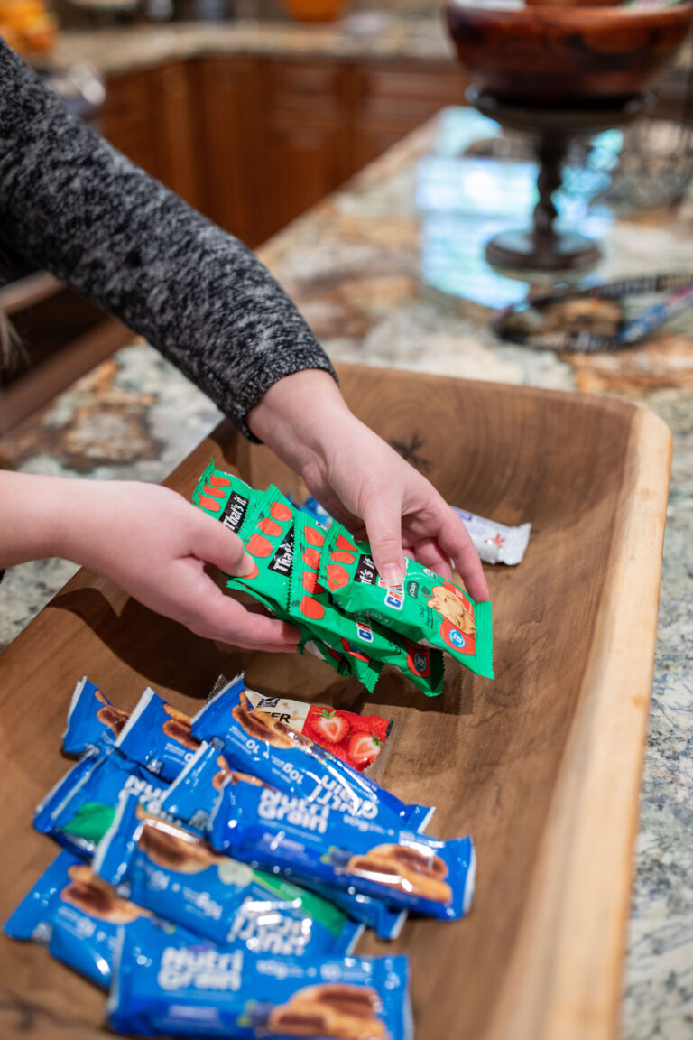 Hands placing snack bars into a wooden tray on a kitchen counter, with blue and green packaged bars.