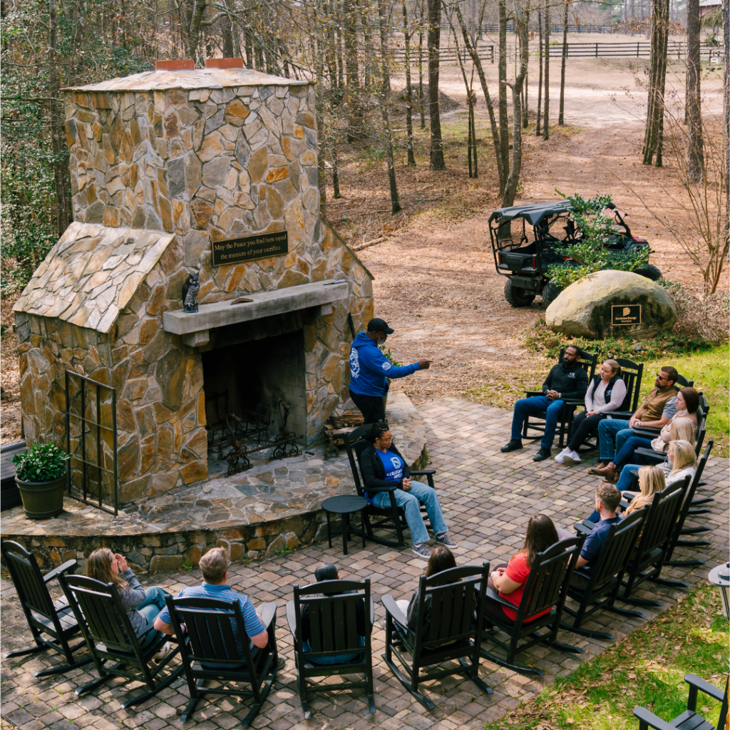 A group of people sit in rocking chairs around an outdoor stone fireplace, listening to a speaker.