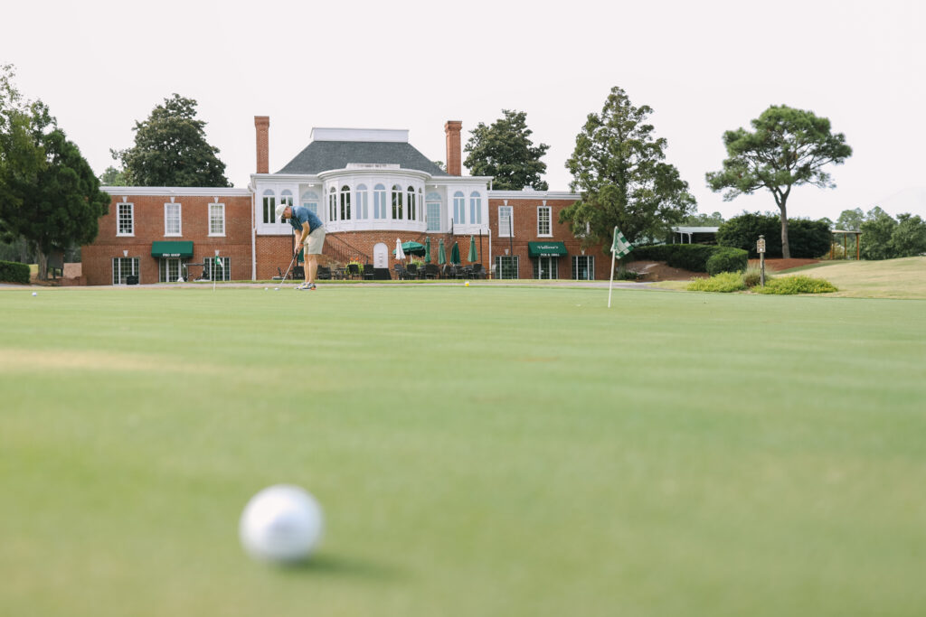 Person putting on a golf green with a clubhouse and trees in the background; a golf ball is in the foreground.