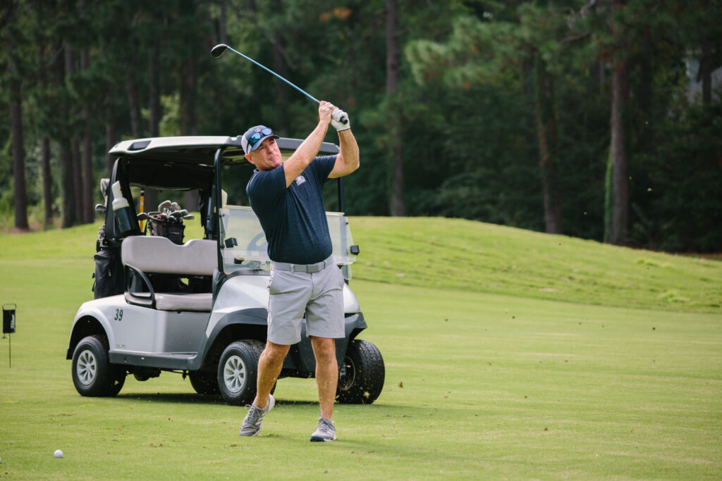 Man swinging a golf club on a golf course with a golf cart in the background.