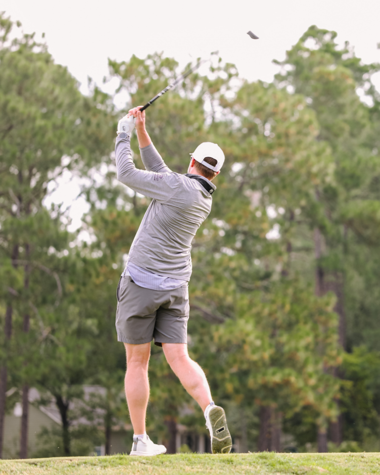 A golfer in a gray outfit swings a club on a grassy course with trees in the background.
