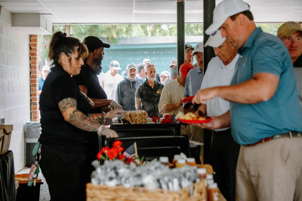 People wait in line as food is served buffet-style at an outdoor event with staff behind a serving table.