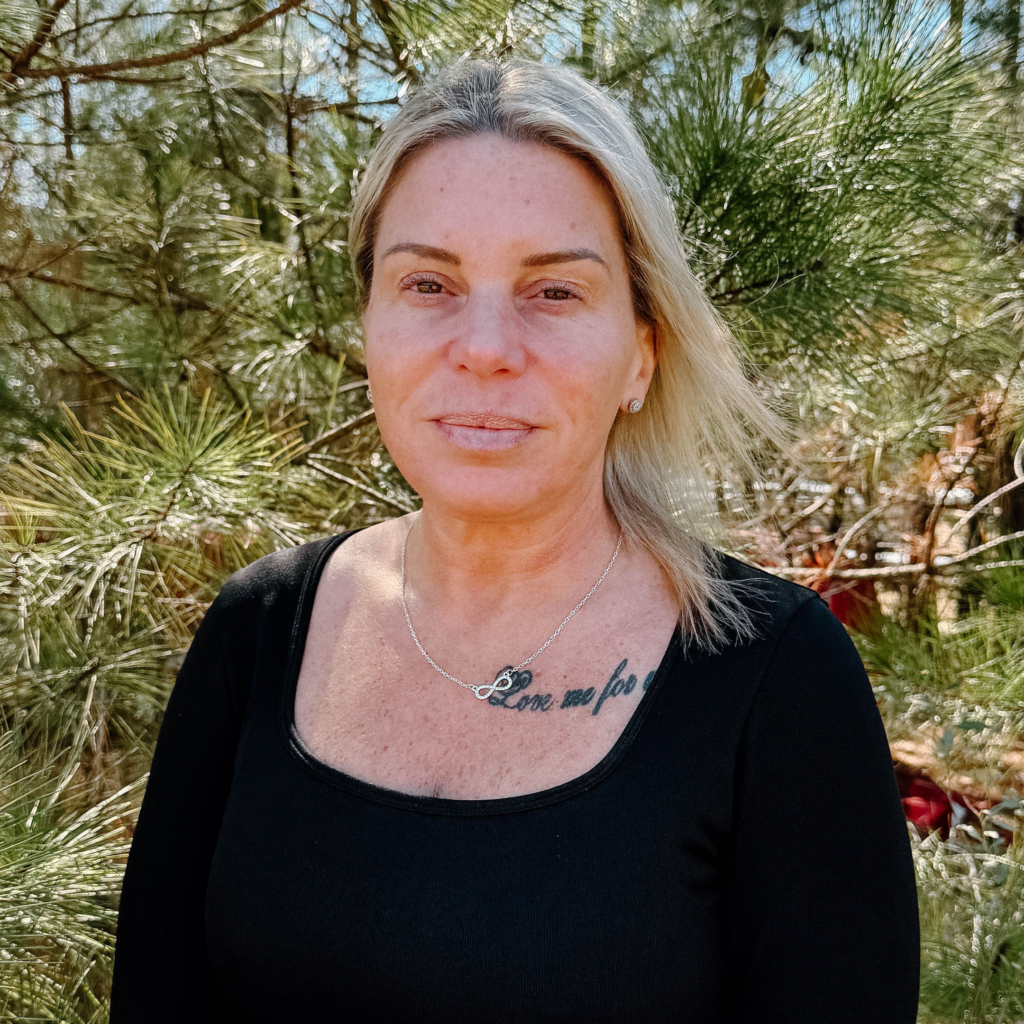A woman in a black top stands outside in front of green pine trees, with wind blowing her blonde hair.
