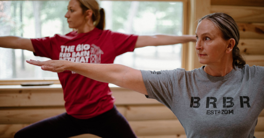 Two women in yoga poses indoors, wearing t-shirts, with wooden walls and a window in the background.